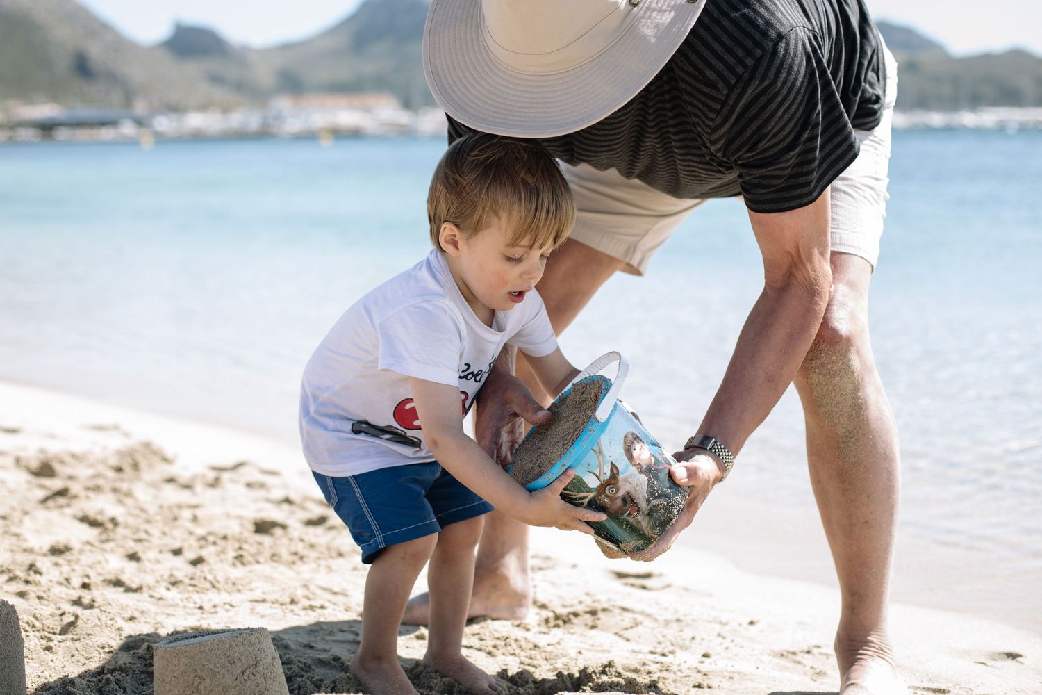 Adult and child explore together on a sunny beach with mountains in the background.