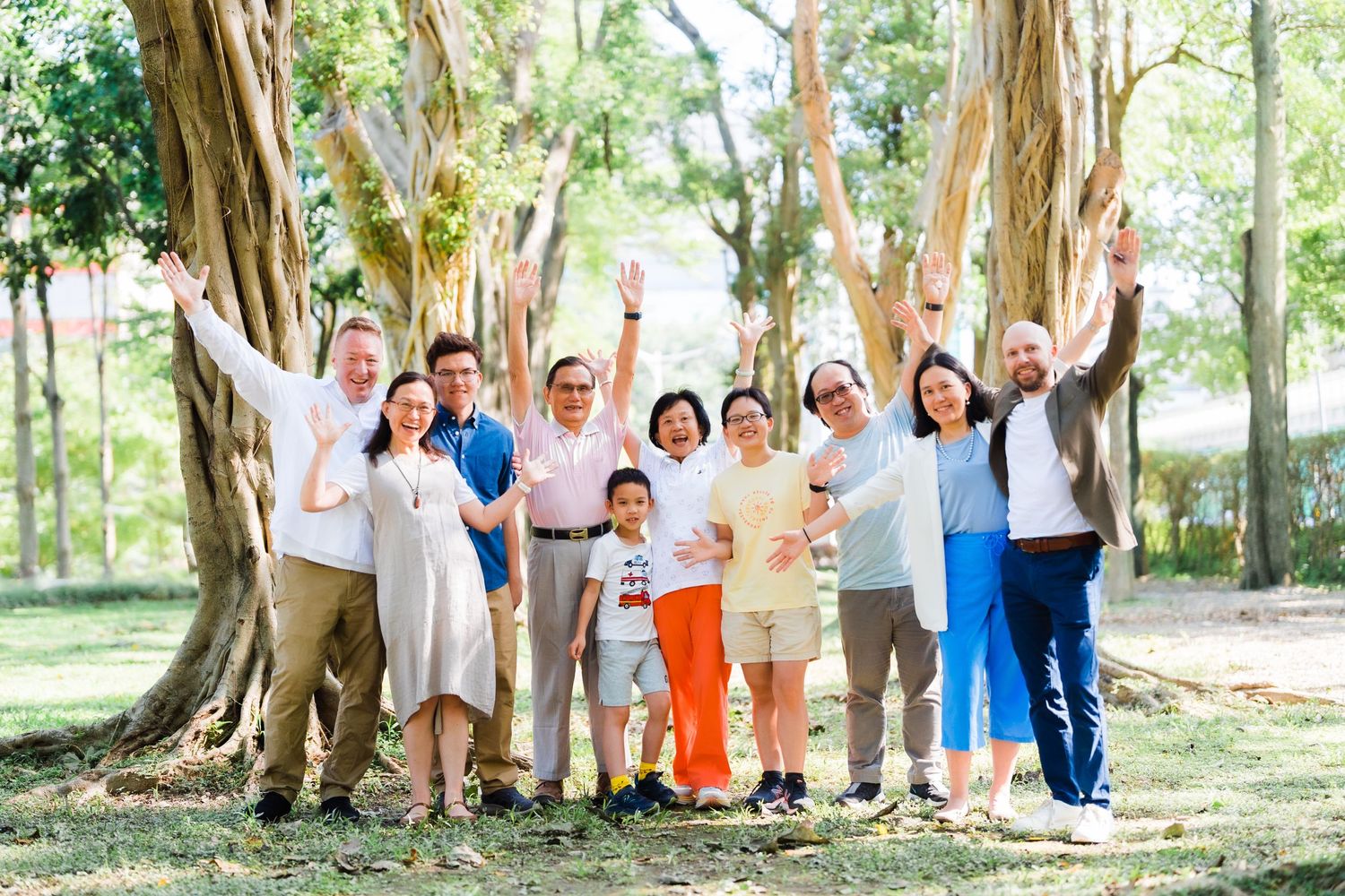 Large family group raises their arms joyfully while posing under large trees in a park.