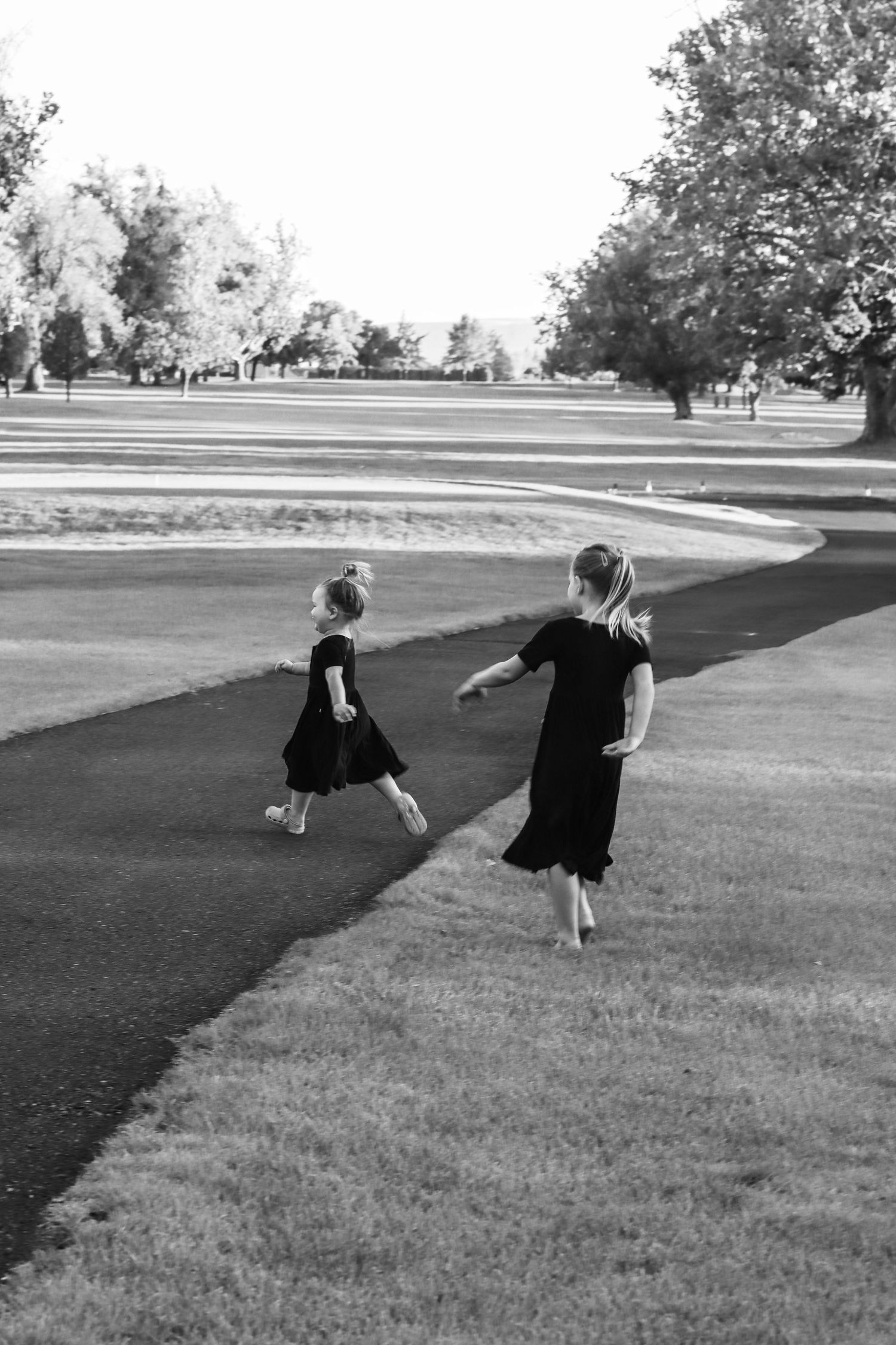 Two young girls in black dresses run barefoot along a winding path in a park, black and white photo.