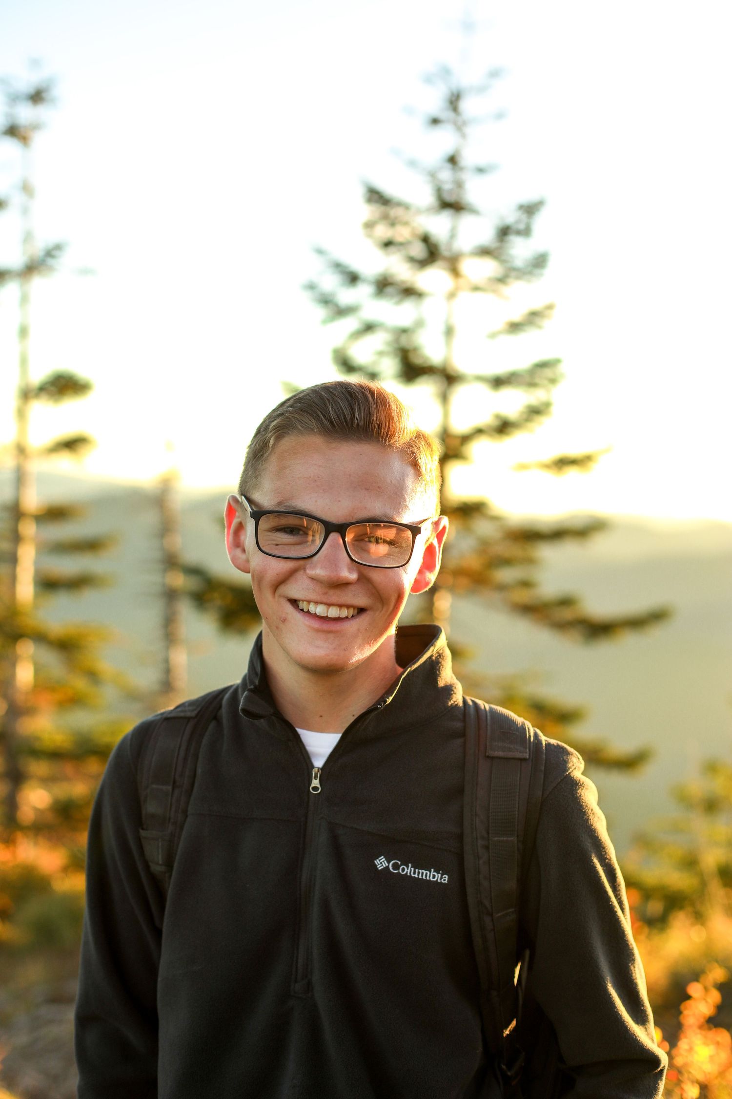 Person in Columbia jacket and glasses smiles during sunset with evergreen trees in background.