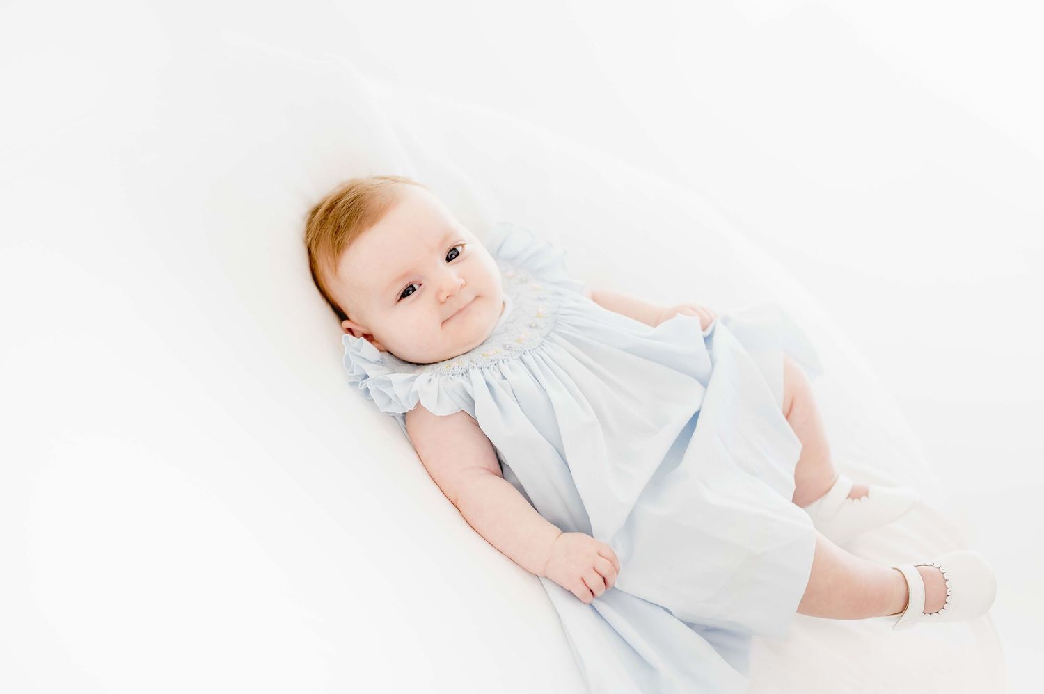 three month old wearing a light blue dress with white shoes taking milestone portraits in an all white studio in houston