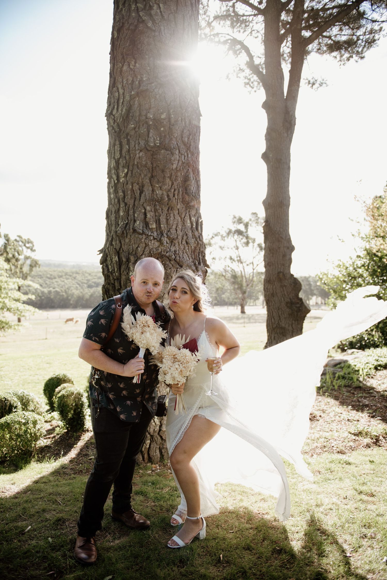 Wedding couple poses playfully by large tree trunk with sunlight streaming through.