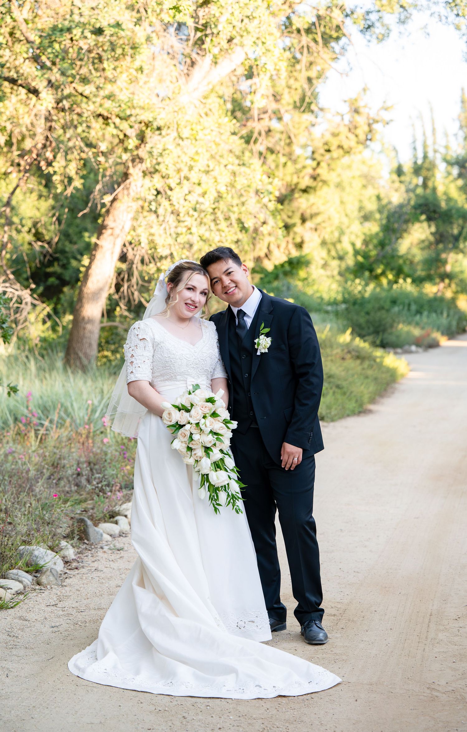 Newlywed couple poses on dirt path with sunlit trees and bride holding cascading bouquet.