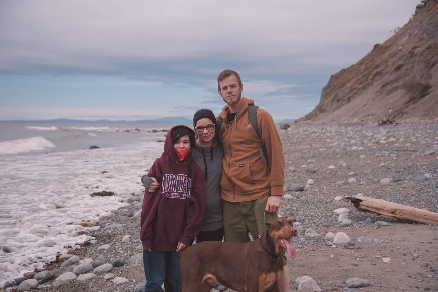 Family of three with a dog posing on a rocky beach with a cliff behind them.