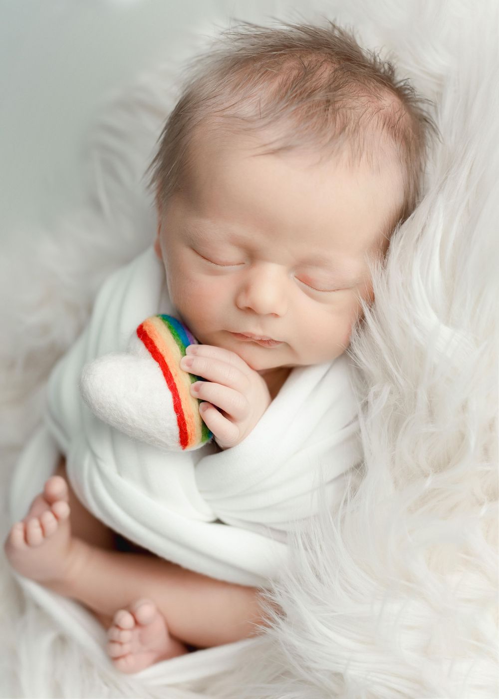 Sleeping newborn baby wrapped in white blanket holding a small rainbow toy. Newborn Photography Batavia Cincinnati Ohio