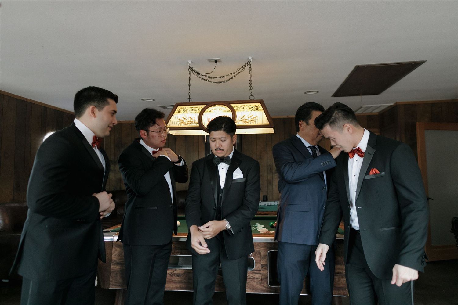 A group of groomsmen in formal attire share a laugh and celebrate together under a vintage ceiling lamp.