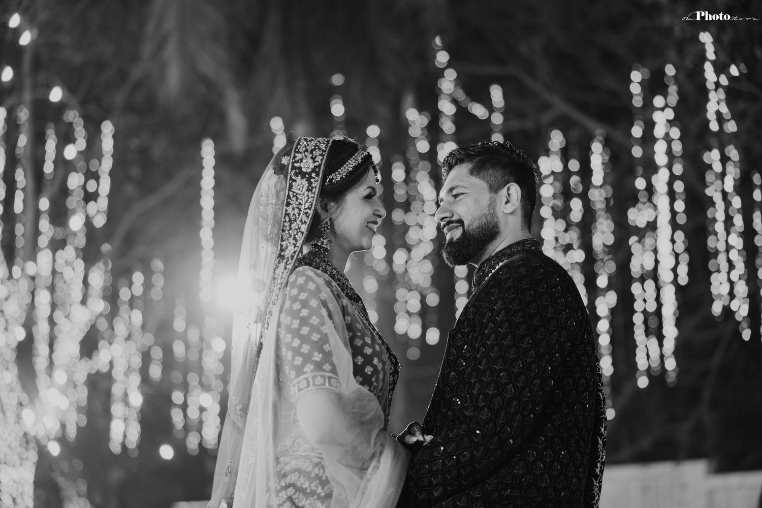 Black and white wedding photo with romantic string light backdrop creating bokeh effect.