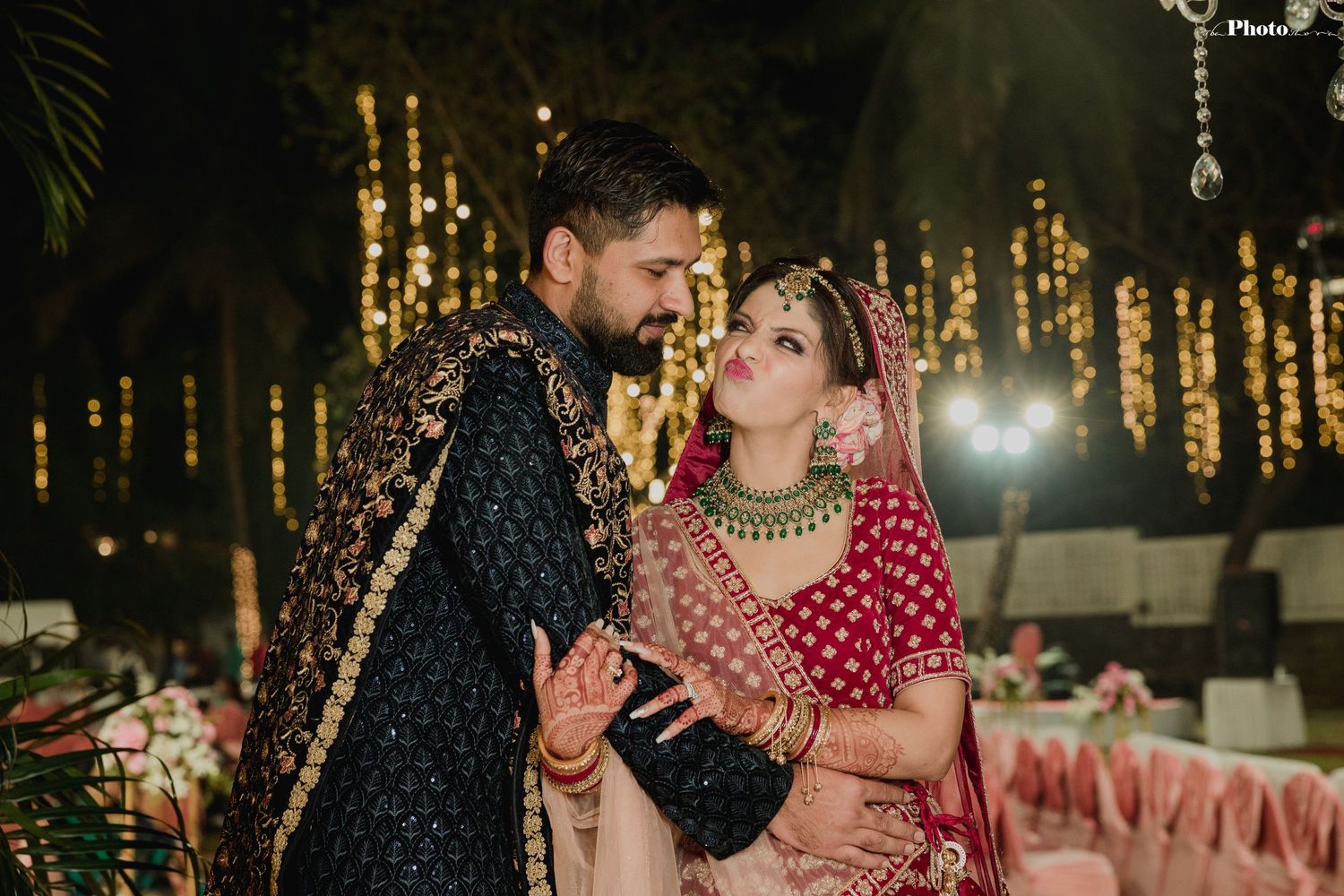 Traditional Indian wedding couple poses against a backdrop of glowing string lights at night.