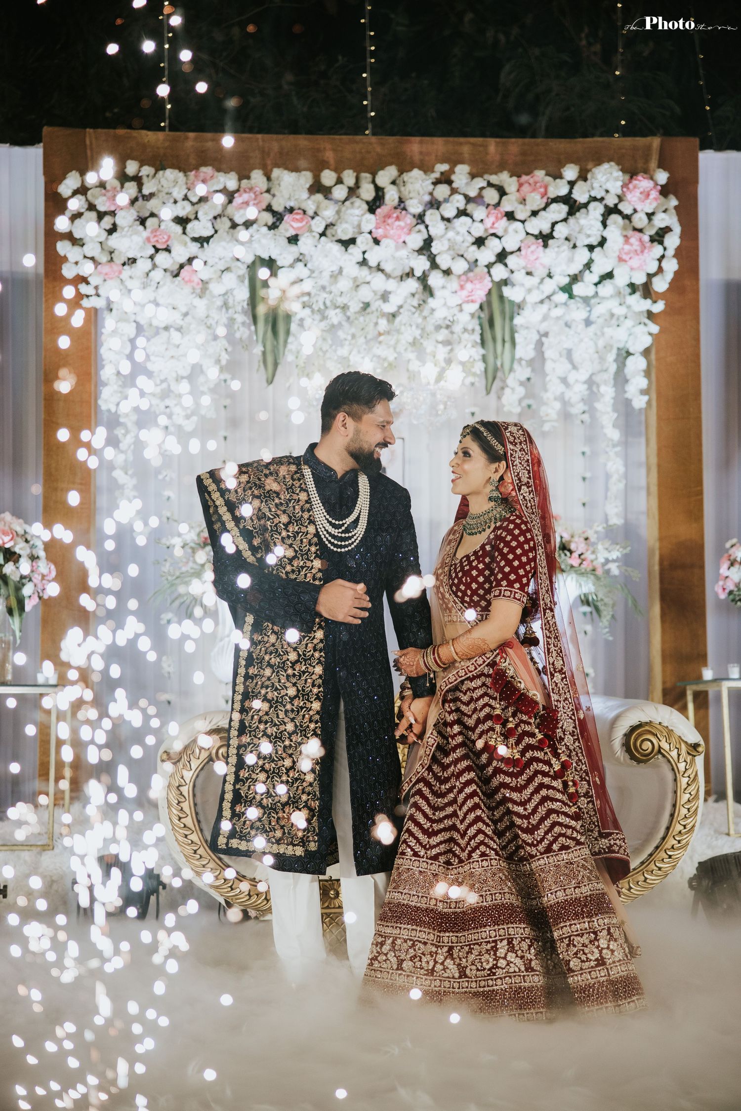 Elegant Indian wedding ceremony setup with white floral backdrop and traditional attire against a romantic lighting display.