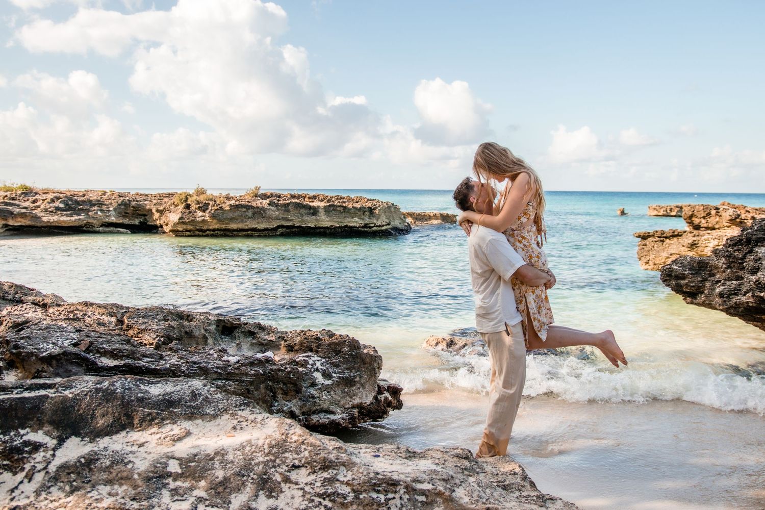 Romantic beach scene with couple embracing on rocky coastline near turquoise waters.