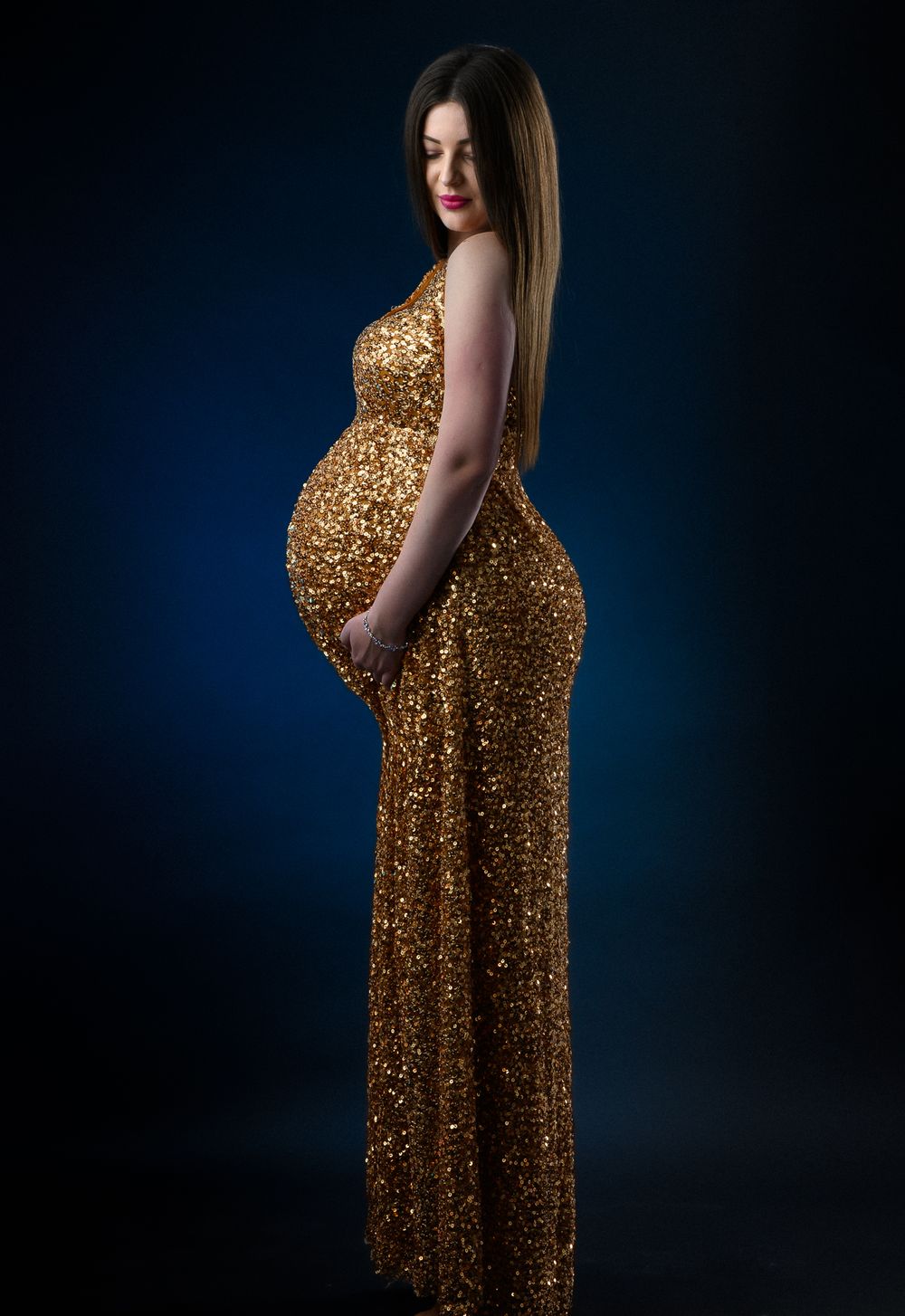 A maternity photoshoot featuring a floor-length gold sequined dress against a dark blue backdrop.