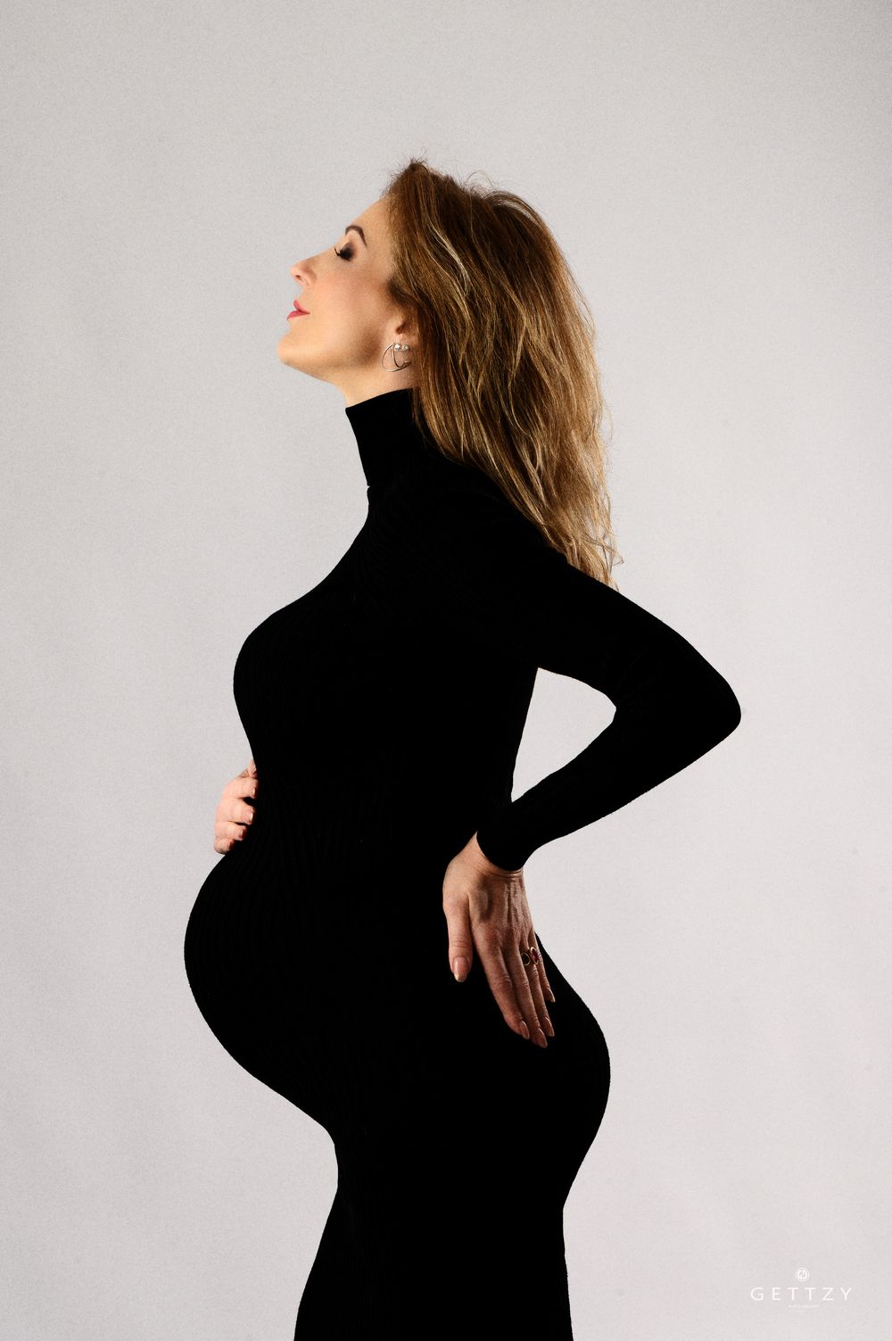 Side profile of an expectant mother in a fitted black turtleneck dress showing her baby bump.