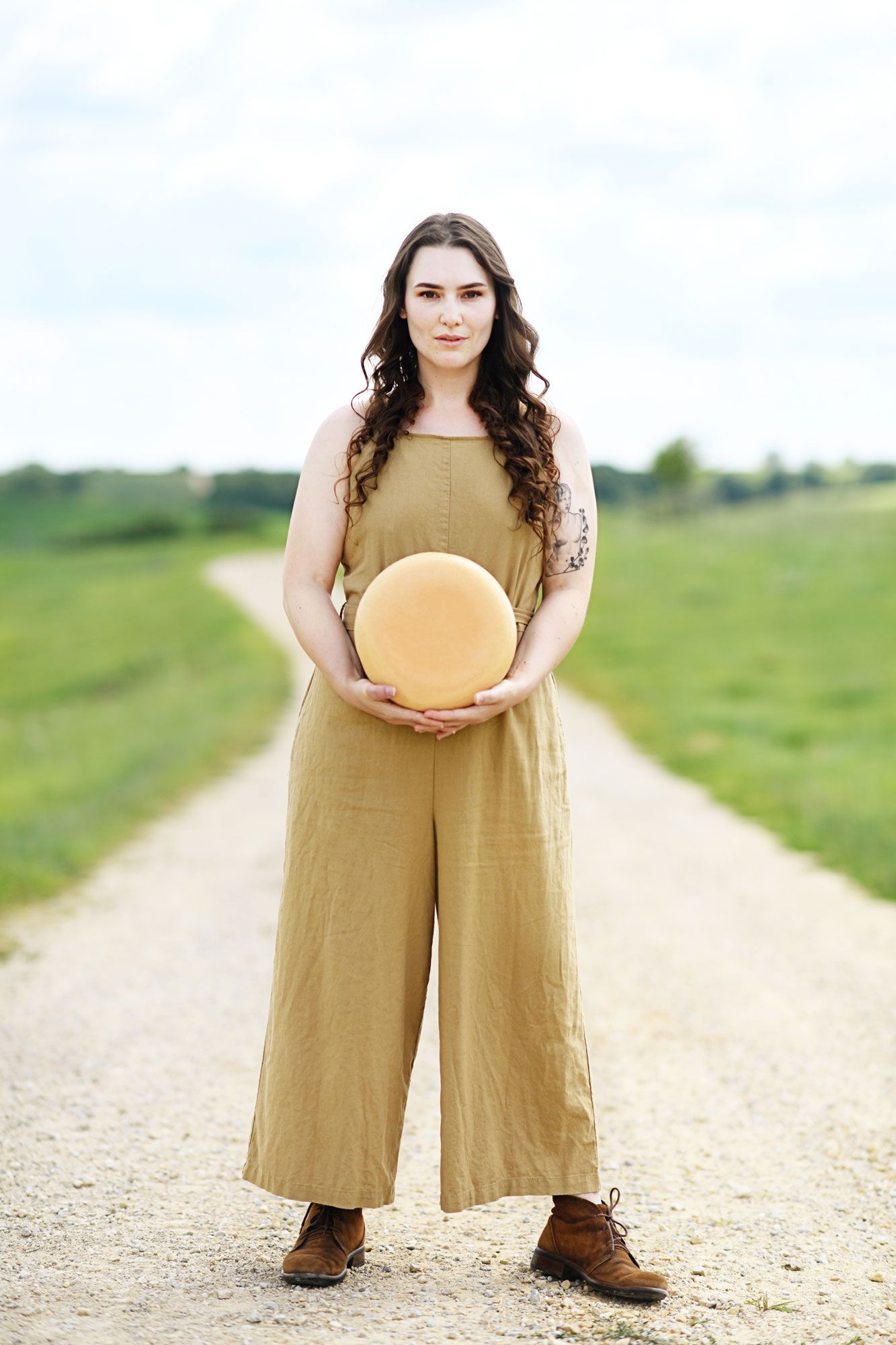 Person in tan jumpsuit standing on dirt road holding round object in rural setting.