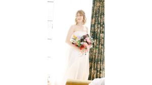 Bride in white gown holding colorful bouquet against floral curtains.