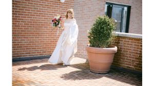 Bride in flowing white dress standing near potted plant on brick patio.