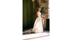 Bride in flowing gown poses against rustic brick wall backdrop.