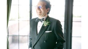 Groom in classic tuxedo and bowtie laughing by window.