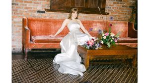 Bride in flowing white gown poses on vintage leather couch with colorful floral arrangement.