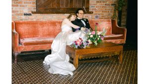 A couple in wedding attire rests on a vintage orange couch with floral arrangements on a wooden coffee table.
