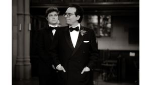 Two people in formal black tuxedos standing in an ornate interior space.