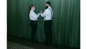 Two people in formal attire adjusting their outfits against a green curtain backdrop.