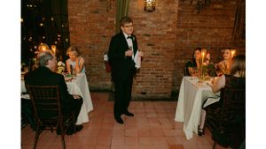 Speaker addressing guests at formal dinner in brick-walled venue with candlelit tables.