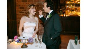 Newlyweds share a joyful moment while cutting their wedding cake at a rustic brick-walled venue.