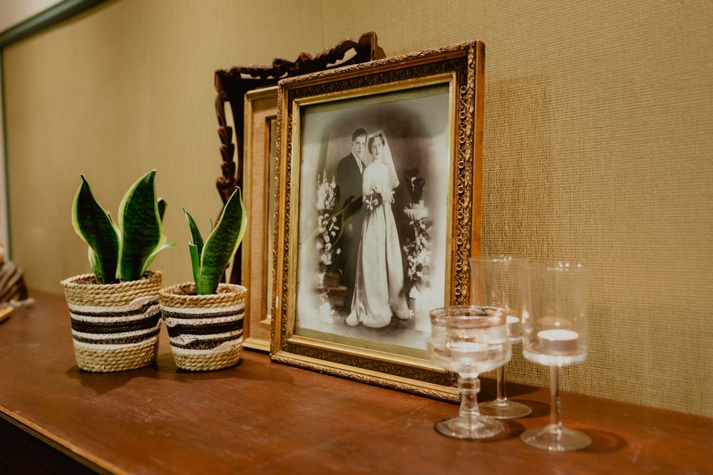Vintage framed photograph displayed on wooden shelf with decorative potted plants and wine glasses.