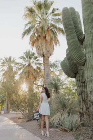 Graduate posing in white dress among desert landscaping and saguaro cacti.
