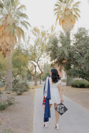 Graduate walking away on campus path with graduation regalia and palm trees in background.
