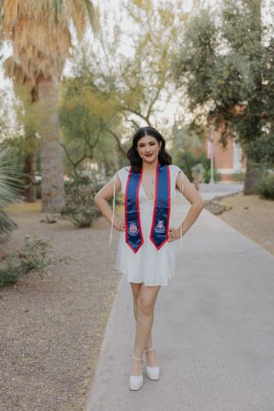 Graduate poses confidently in white dress with blue and red graduation stoles on campus path.