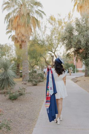 Graduate in academic regalia walking along desert pathway with palm trees.