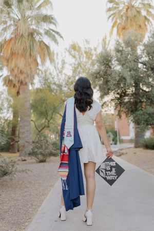 Graduate in white dress and graduation stoles walks on palm tree-lined path carrying graduation cap.