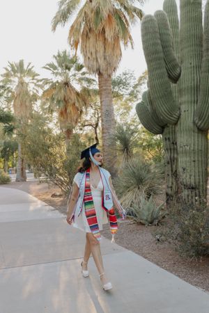 Graduate walking along desert path surrounded by saguaro cacti and palm trees.