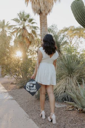 Graduate walking through desert garden path in white dress and graduation cap.