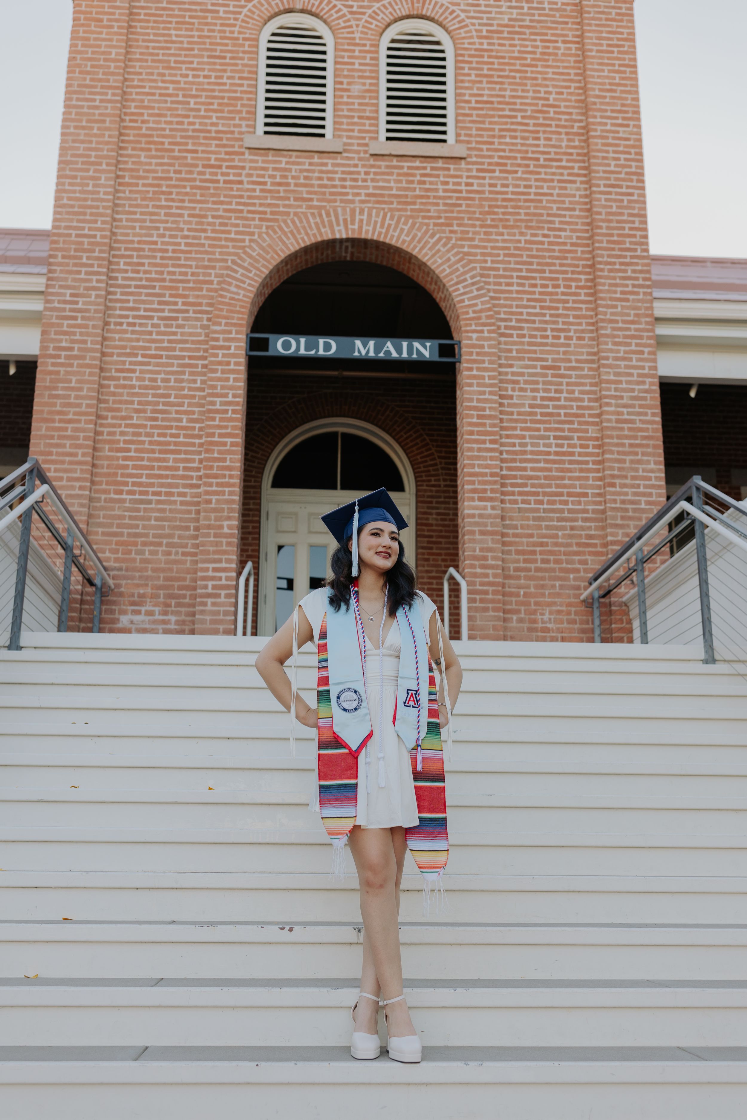 Graduate in academic regalia poses on brick building steps during graduation photoshoot.