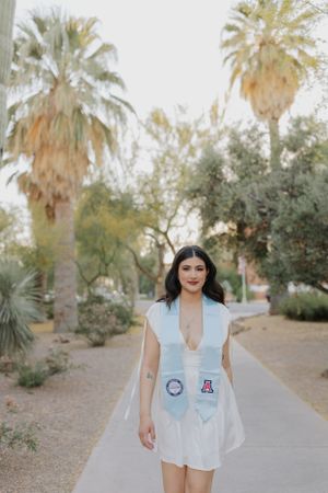 Graduate in white dress poses with graduation stoles on palm tree-lined campus walkway.