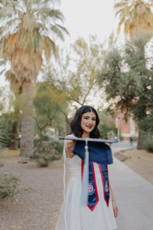 Graduate holds up graduation stoles while posing on campus walkway with palm trees.