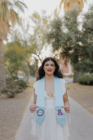 Graduate wearing white dress and light blue graduation stoles smiles on campus path.