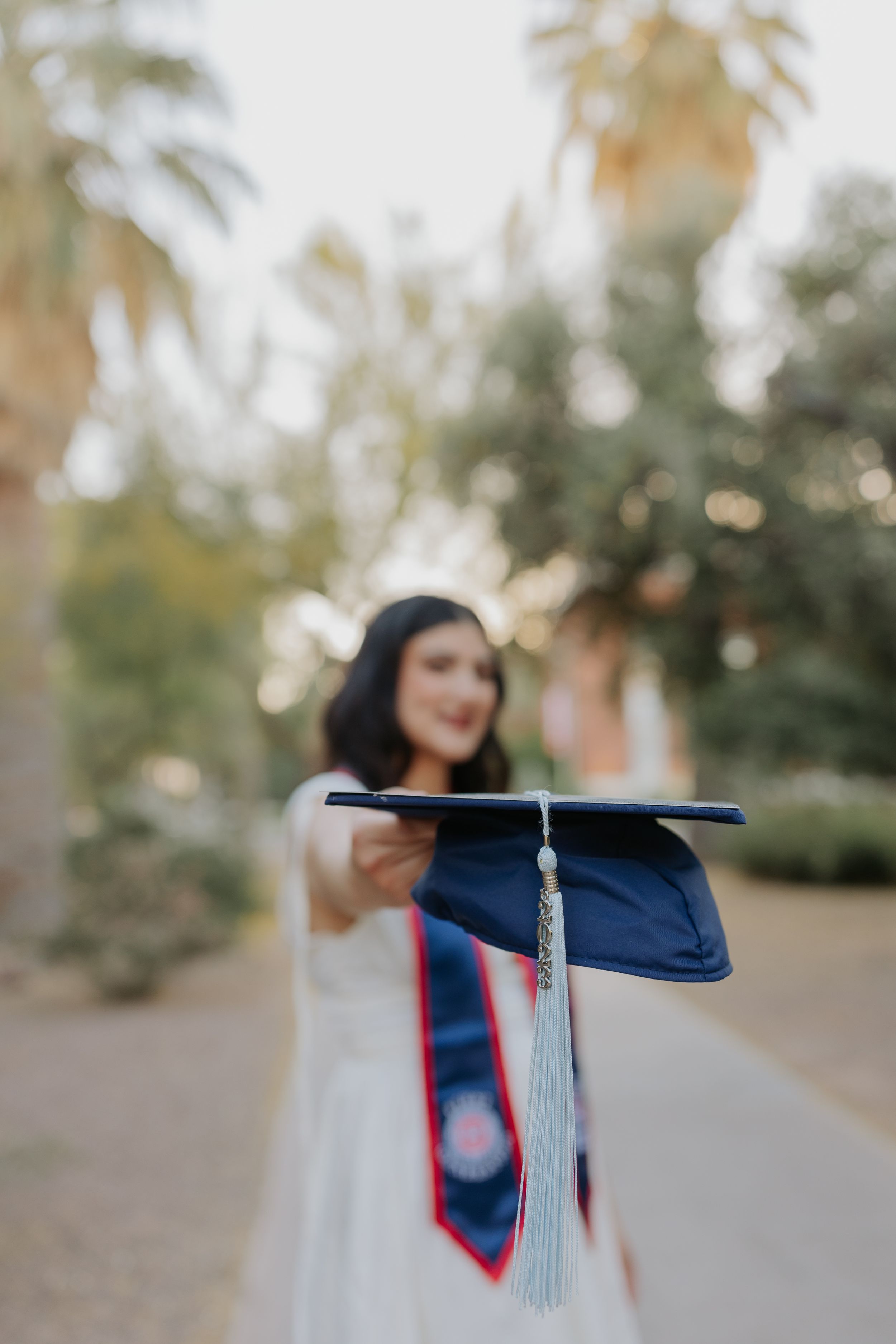 Graduate holds up blue graduation cap with tassel against blurred palm tree background.