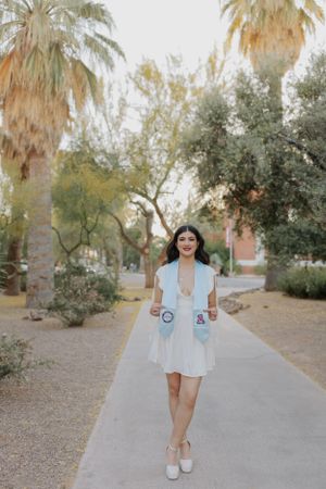 Graduate in white dress and graduation stoles poses on palm-lined walkway.
