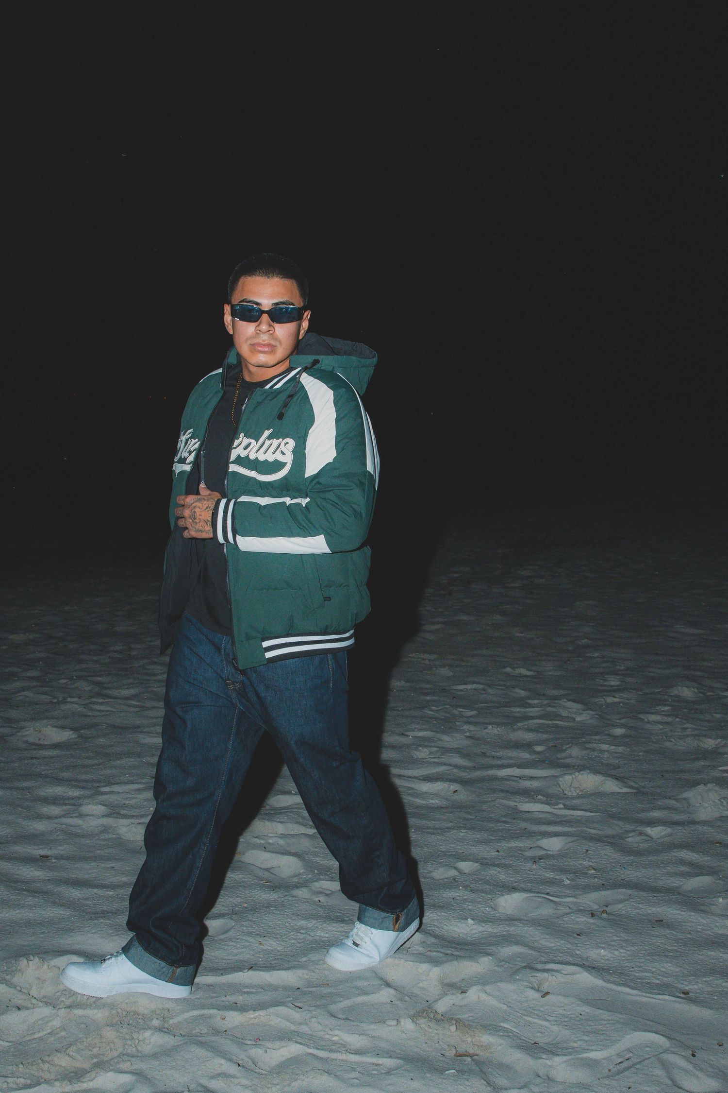 Person wearing a green sports jacket and baggy jeans posing on a beach at night.