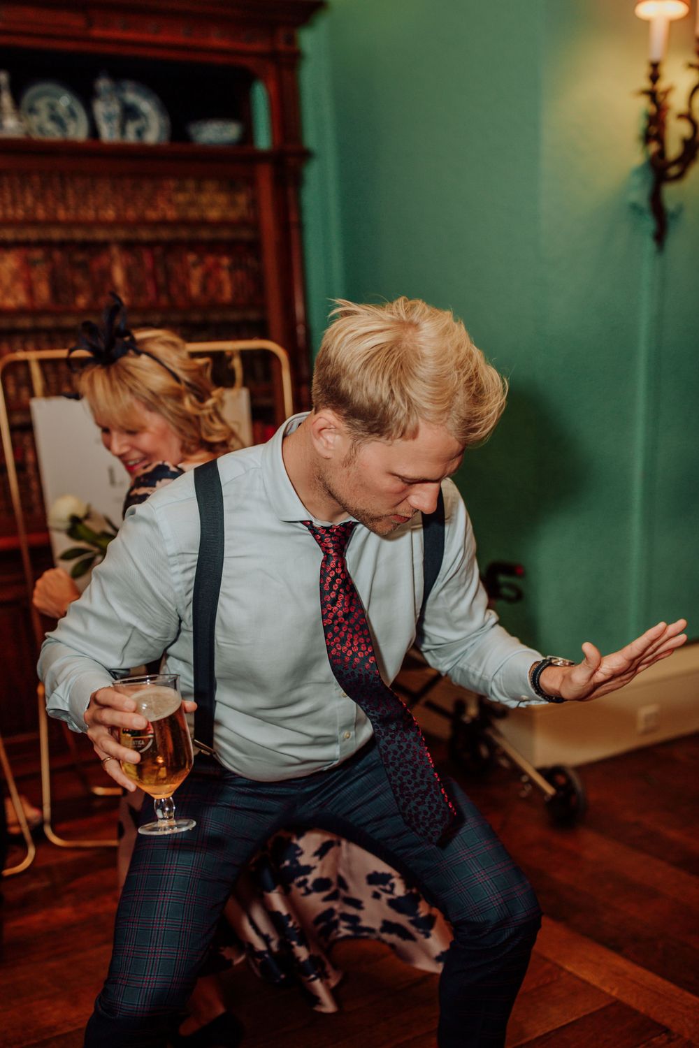 Wedding guest dances enthusiastically in suspenders and tie at indoor reception.