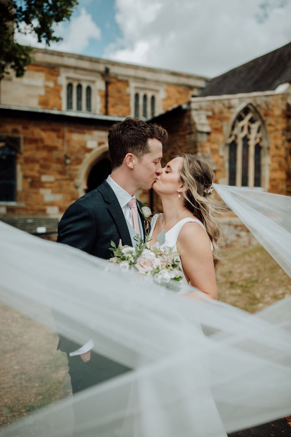A romantic wedding kiss captured in front of a historic stone church with flowing veil.