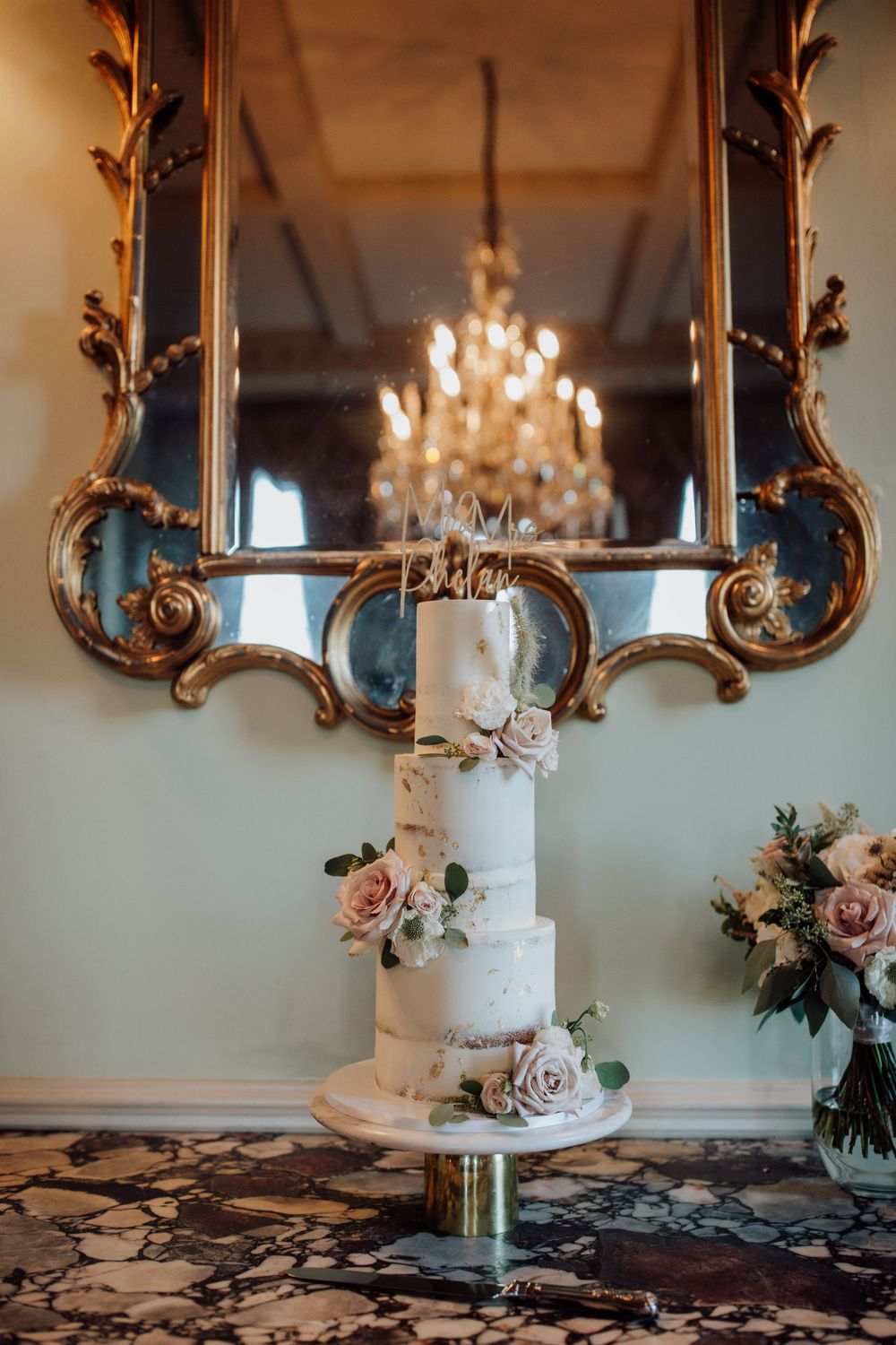 Elegant white wedding cake with blush roses displayed in front of an ornate gold mirror and crystal chandelier.