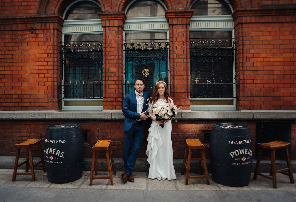 Newlyweds seated on wooden stools outside a rustic brick building with arched windows.