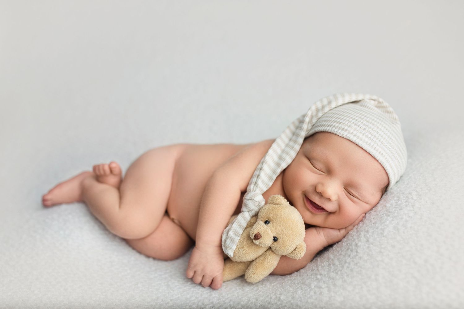 Sleeping newborn wearing white cap posed with small teddy bear on light backdrop.