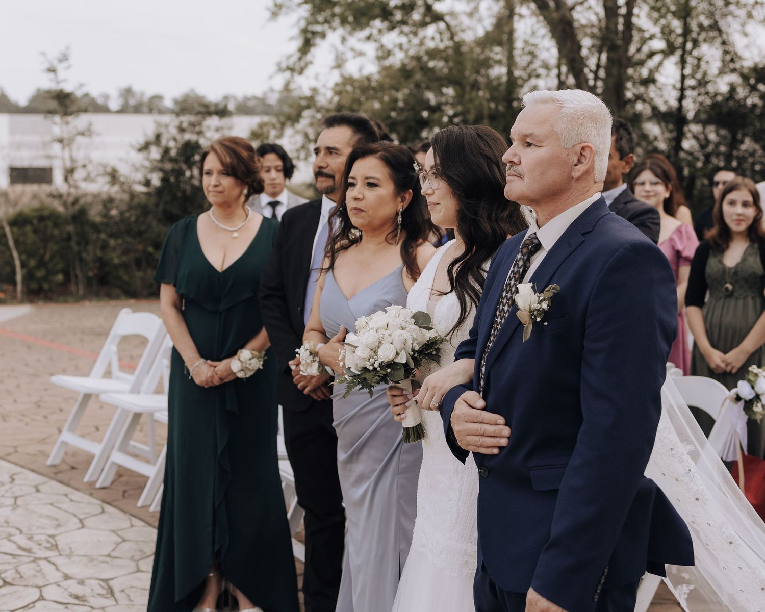 Wedding guests in formal attire stand together during an outdoor ceremony while watching the proceedings.