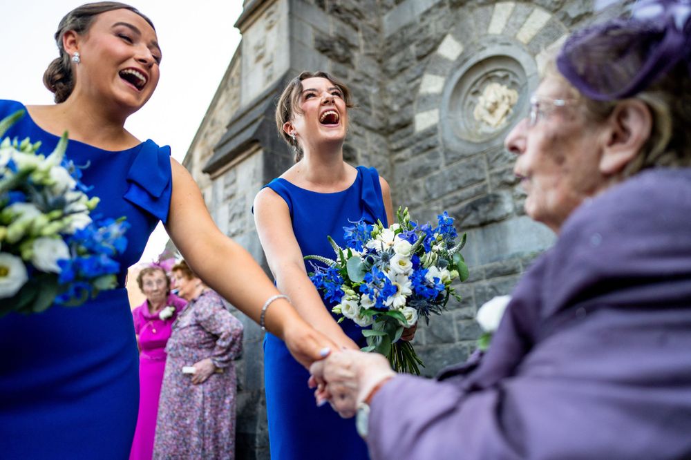 Bridesmaids in royal blue dresses holding white and blue bouquets share a joyful moment outside a church.