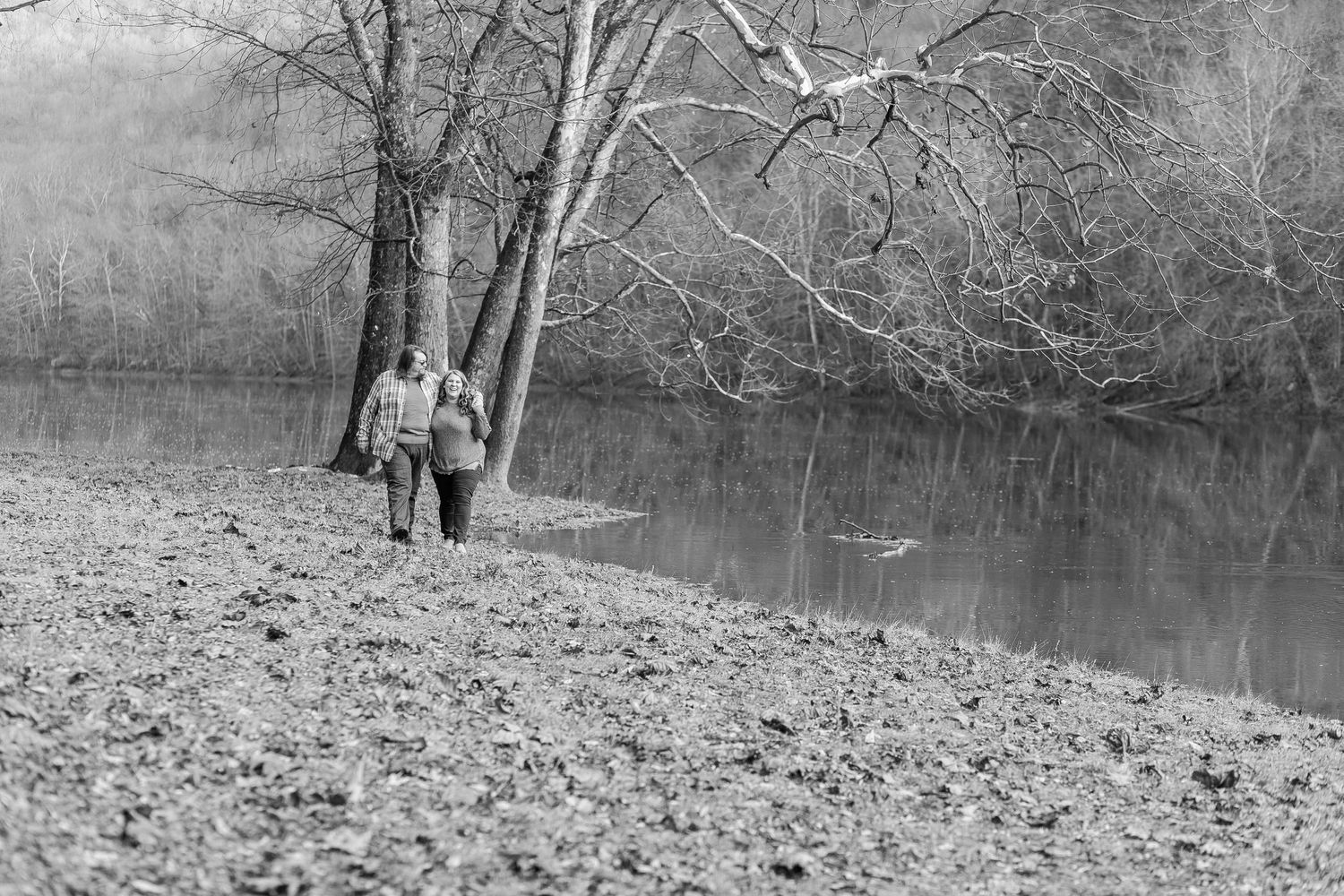 A black and white photo of two people standing by a tranquil lake with bare trees.